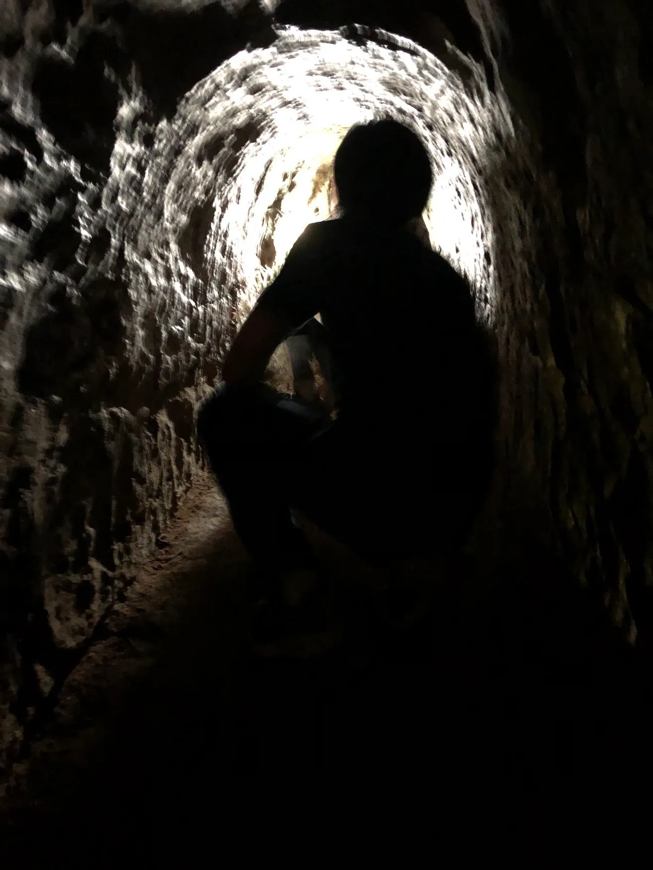 A crouching figure silhouetted against daylight at the far end of a narrow earth tunnel