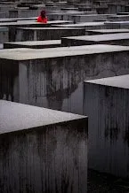 Rows of grey concrete slabs receding into the distance, a single visitor in a red coat among them
