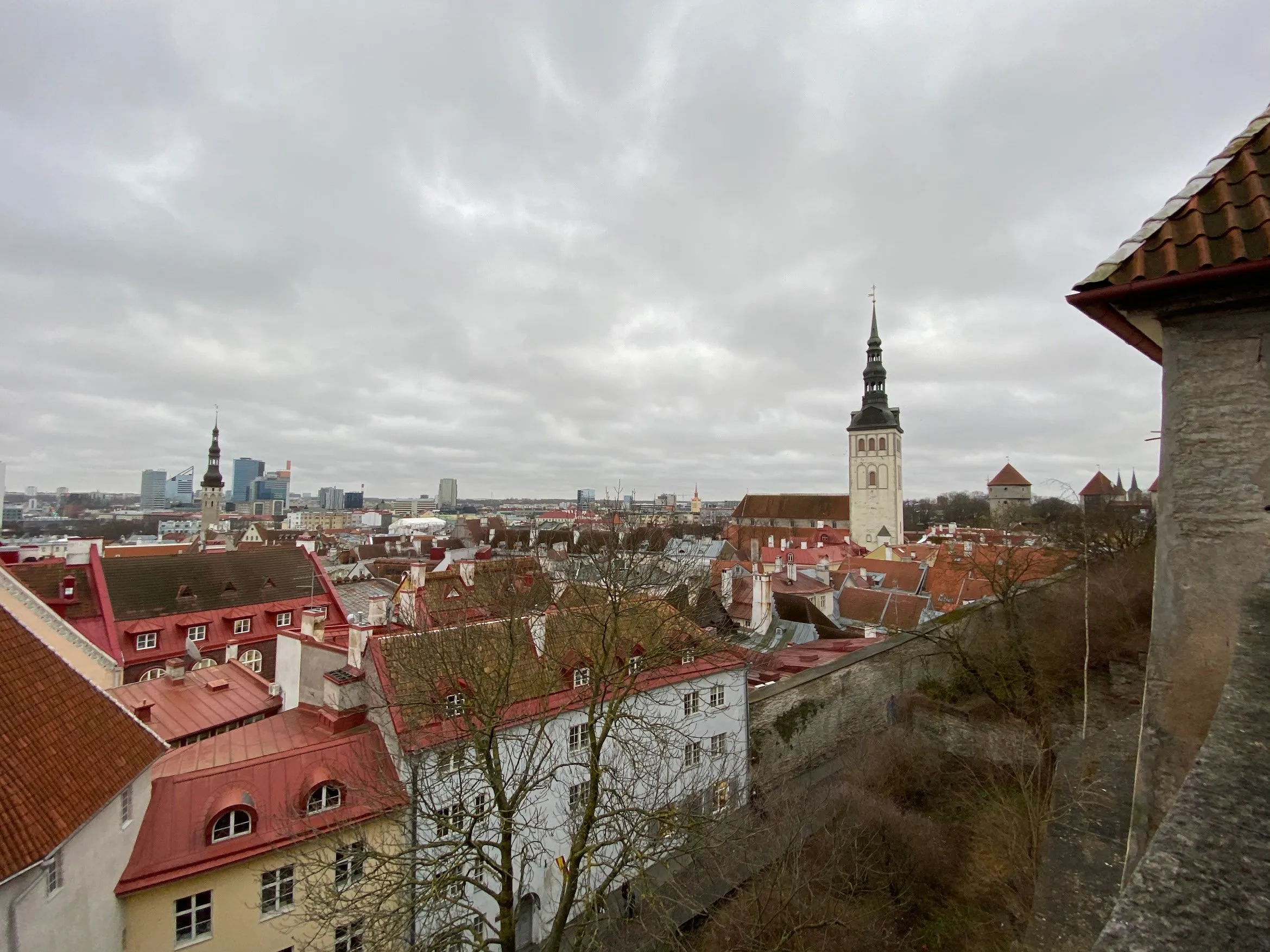 Red-tiled roofs and a church spire in Tallinn’s old town under heavy cloud, modern towers on the horizon