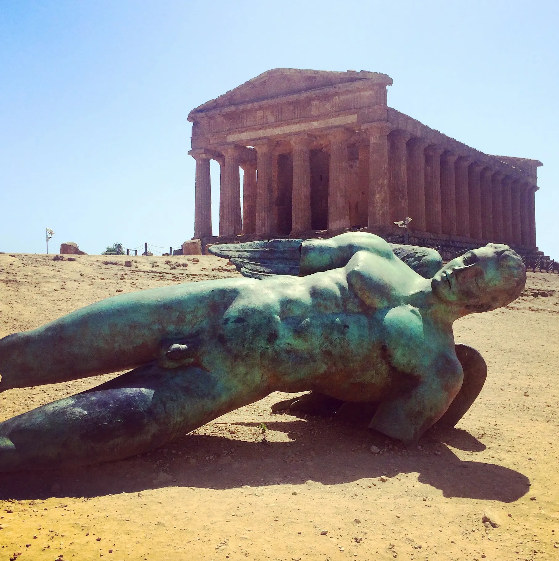 A large bronze figure with one wing lying on the sand before the columned Greek temple