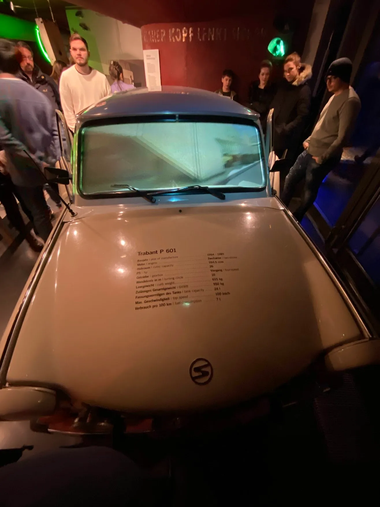 A pale Trabant on display in a dim museum hall, visitors standing around it