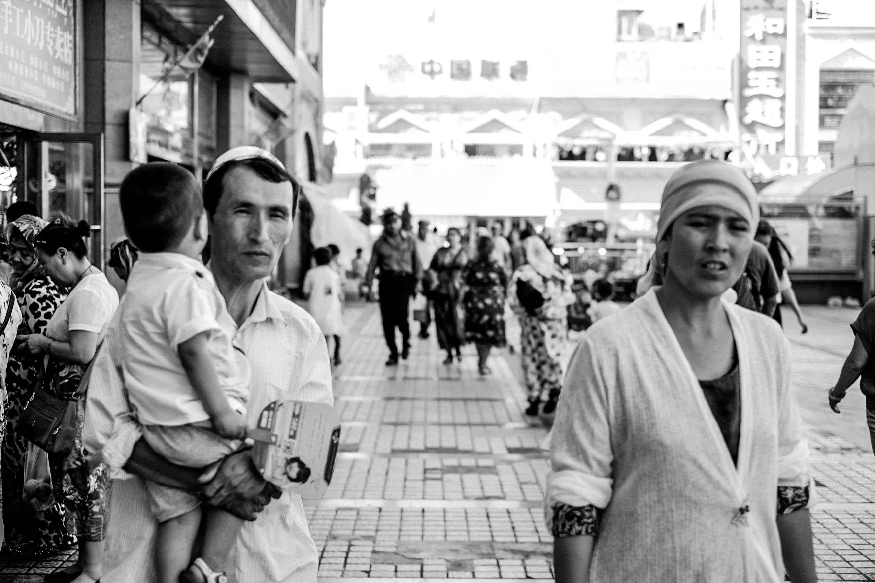 A black and white street scene: a man carrying a small child past market stalls, a woman in a headscarf in the foreground